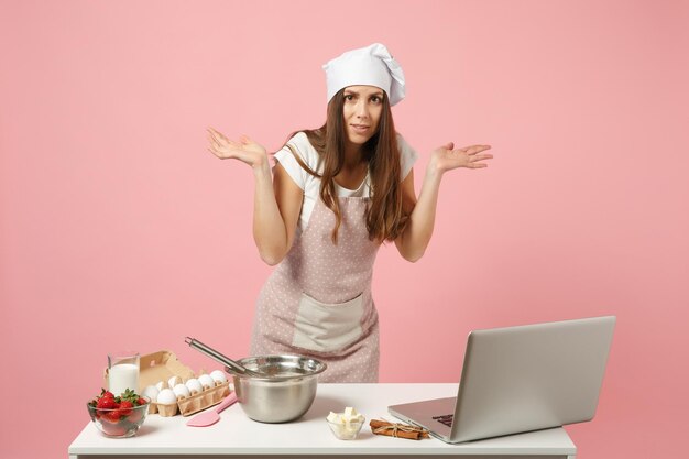 Szef kuchni kucharz cukiernik lub piekarz w fartuch biały t-shirt, kapelusz kucharzy toczek gotowania świąteczny tort lub ciastko przy stole, szukając przepisu na laptopie pc na białym tle na różowym pastelowym tle. Makieta koncepcji.