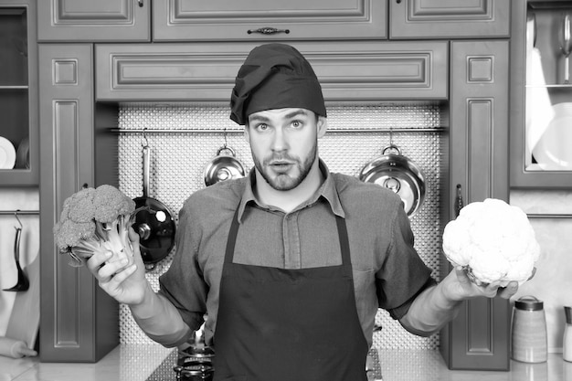 Szef kuchni macho ze zdziwioną twarzą trzyma kalafior i brokuły