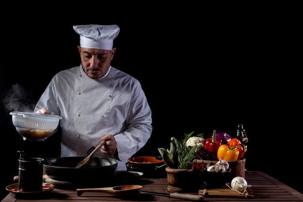 Szef Kuchni Mężczyzna W Białym Mundurze Wlewając Gotowane Spaghetti Do Woka Do Gotowania Makaronu Z Warzywami. Za Kulisami Przygotowywania Tradycyjnej Włoskiej Potrawy Na Czarnym Tle