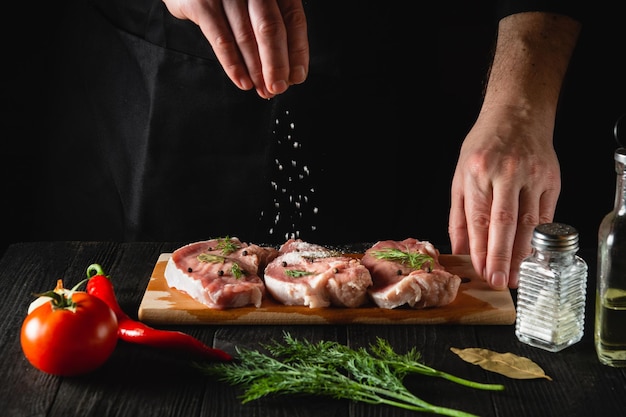 Szef kuchni posypuje mięso solą. Przygotowanie mięsa przed pieczeniem. Środowisko pracy w kuchni restauracji lub kawiarni
