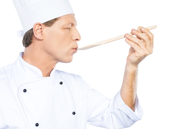 Zdjęcie szef kuchni próbuje posiłek. widok z boku pewnego siebie dojrzałego szefa kuchni w białym mundurze, który ma zamknięte oczy, próbując jeść z drewnianej łyżki i stojąc na białym tle