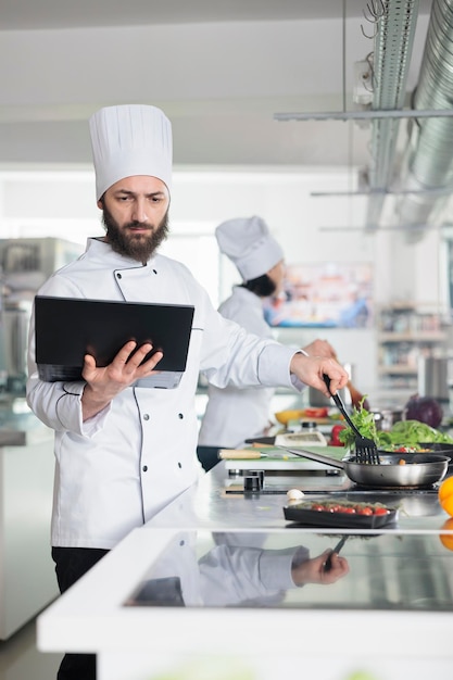 Szef Kuchni Przygotowuje świeży Organiczny Dodatek Warzywny Za Pomocą Patelni I łopatki, Patrząc Na Ekran Laptopa. Główny Kucharz Z Komputerową Burzą Mózgów, Przygotowujący Przepisy Na Dania Dla Smakoszy Na Konkurs Kulinarny, Który Odbywa Się W Restauracji.