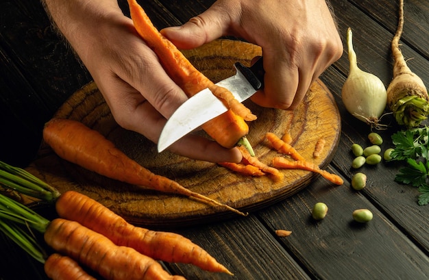 Szef kuchni rękami noża łuszczy marchewki na kuchennej desce do cięcia, aby przygotować posiłek warzywny Koncepcja jedzenia wegetariańskiego