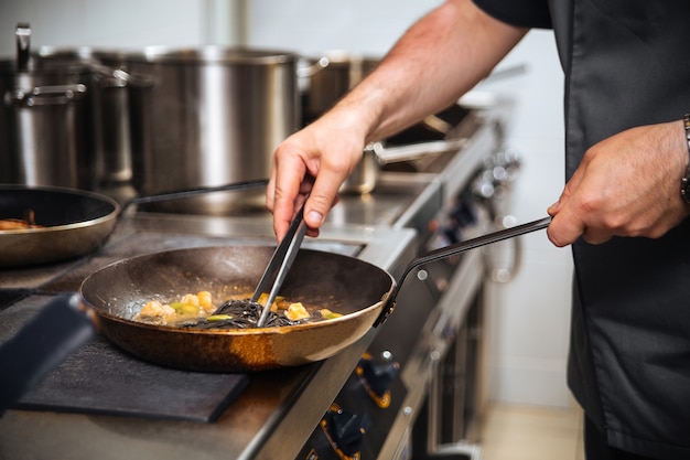 Szef kuchni restauracji gotuje czarne spaghetti na patelni ze szczypcami