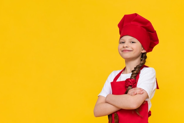 Szef kuchni to mała dziewczynka w czerwonym kapeluszu i fartuchu z założonymi rękami na piersi Miejsce na reklamę na żółtym, odizolowanym tle Miejsce na kopię