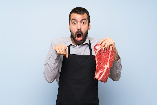 Zdjęcie szef kuchni trzymając surowe mięso zaskoczony i wskazując przód