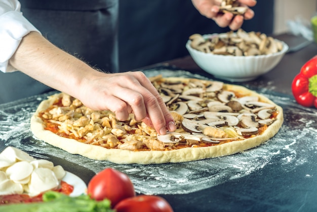 Szef kuchni w czarnym fartuchu stawiający pieczarki na pizzy