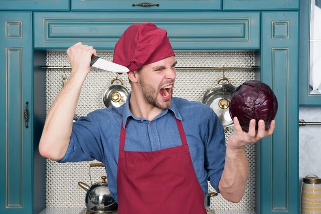 Szef kuchni z nożem krzyczy na fioletową kapustę Zły człowiek w czerwonym kapeluszu i fartuchu w kuchni Wegetariańskie menu i zdrowa dieta Przygotowanie żywności i przepisy kulinarne Warzywa i przybory kuchenne do gotowania potraw