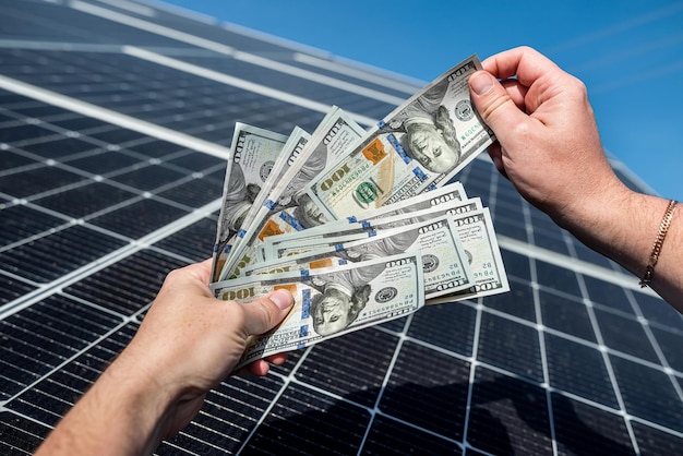 Szef w kurtce z młodymi rękami trzymający dużą ilość dolarów na tle paneli słonecznych Koncepcja zielonej energii elektrycznej