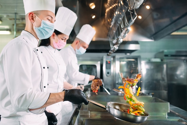 Szefowie kuchni w maskach ochronnych i rękawiczkach przygotowują jedzenie w kuchni restauracji lub hotelu.