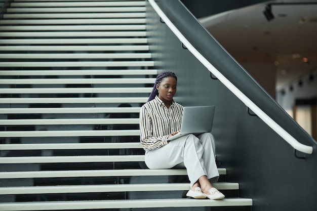 Szeroki kąt portret afroamerykańskiej bizneswoman w czarno-białym stroju za pomocą laptopa siedząc na schodach w biurze, kopia przestrzeń