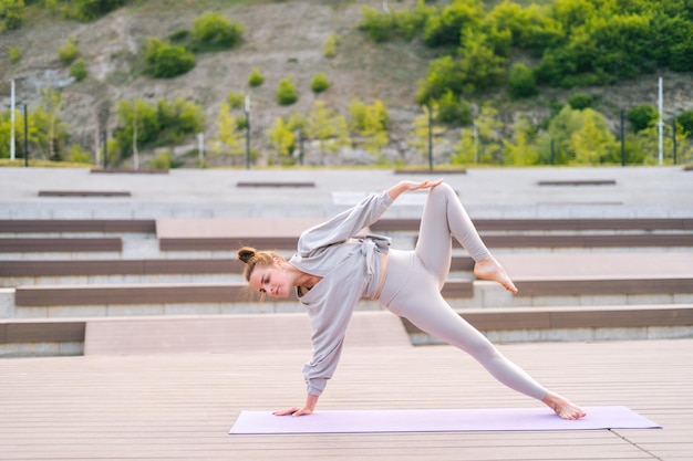 Szerokie ujęcie elastycznej młodej joginki robi jogi stojącej na macie fitness w letni dzień na zewnątrz Jogini pasuje sportowa kobieta ubrana w odzież sportową robi ćwiczenia rozciągające na zewnątrz