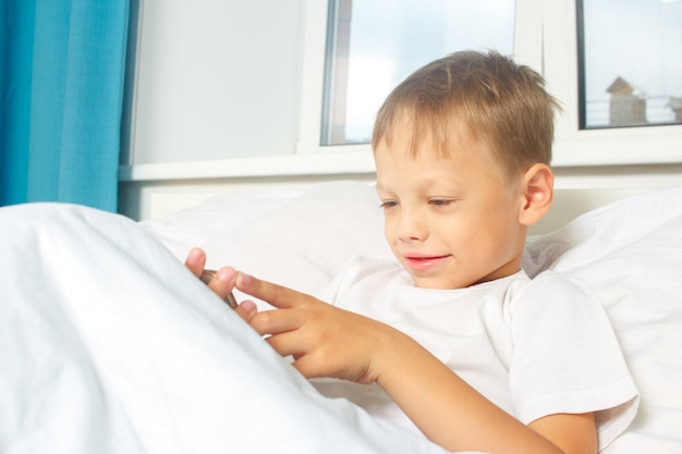Sześcioletni chłopiec odpoczywa, leżąc w łóżku i trzymając w rękach smartfona. Dziecko z gadżetem w domu.