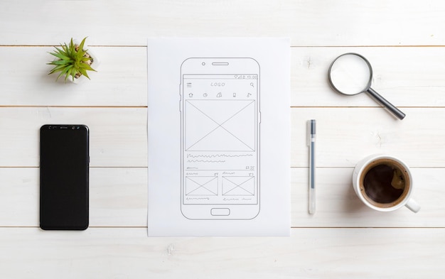 Szkic aplikacji mobilnej na papierze Widok z góry na biurko projektanta stron internetowych Koncepcja projektowania interfejsu użytkownika Inteligentny telefon obok
