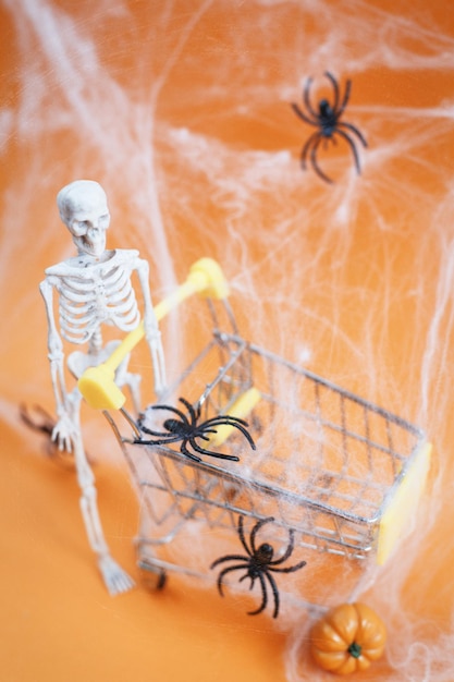 Szkielet z dynią w pobliżu wózka pajęczyny i pająki na pomarańczowym tle Koncepcja zakupów na Halloween
