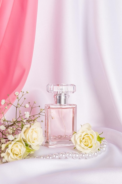Szklana butelka perfum na białym satenowym tle z różami i perłami Modna koncepcja naturalnych zapachów Widok pionowy
