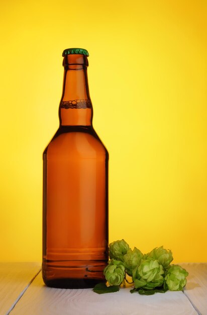 Szklana butelka świeżego piwa z zielonych szyszek chmielowych na jasnym drewnianym stole na żółtym tle.