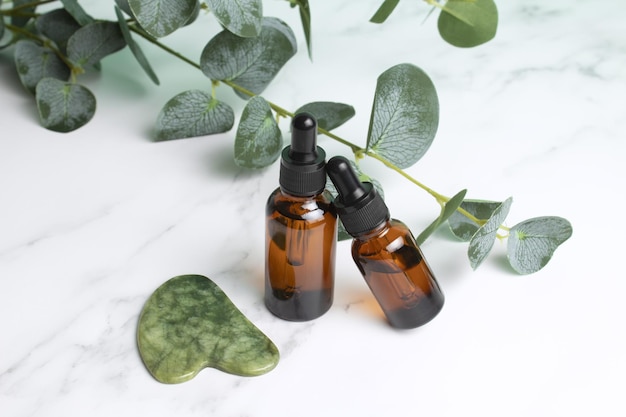 Szklane butelki z olejowym kamieniem Gua sha do masażu twarzy na marmurowym tle z liśćmi eukaliptusa Kosmetyczna pielęgnacja skóry twarzy i spa Naturalna koncepcja leczenia