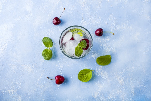 Szklanka letniej lemoniady lub mrożonej herbaty. Odświeżający chłodny napój z wiśnią i miętą na niebieskim stole. Koktajl Mojito z kostkami lodu.