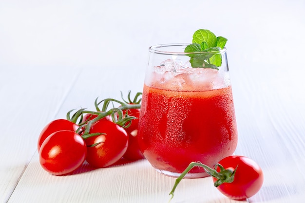 Szklanka soku pomidorowego, kostki lodu i gałązka mięty na białym drewnianym stole. Pojęcie zdrowej żywności wegetariańskiej.