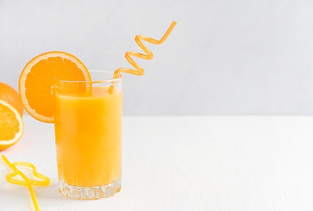 Szklanka świeżego soku pomarańczowego podana ze słomą i plasterkiem owoców cytrusowych na białym drewnianym tle