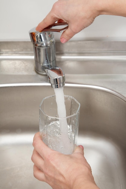 Szklanka w kobiecej dłoni jest wypełniona czystą przezroczystą wodą pod kranem z metalowym zlewem w tle