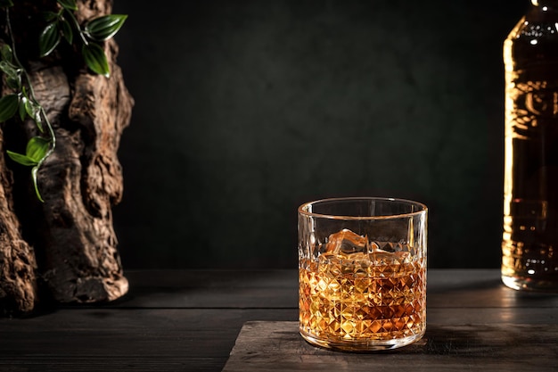 Szklanka whisky z lodem i butelką na drewnianym stole na ciemnym tle z miejsca na kopię