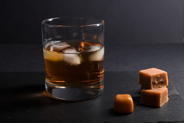 Szklanka whisky z lodem i karmelowymi cukierkami na czarnej kamiennej tabliczce. Widok z boku
