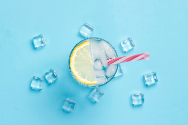Szklankę wody lub napój z lodem i cytryną na niebieskiej powierzchni z kostkami lodu. Pojęcie gorącego lata, alkohol, napój chłodzący, gasi pragnienie. Leżał płasko, widok z góry