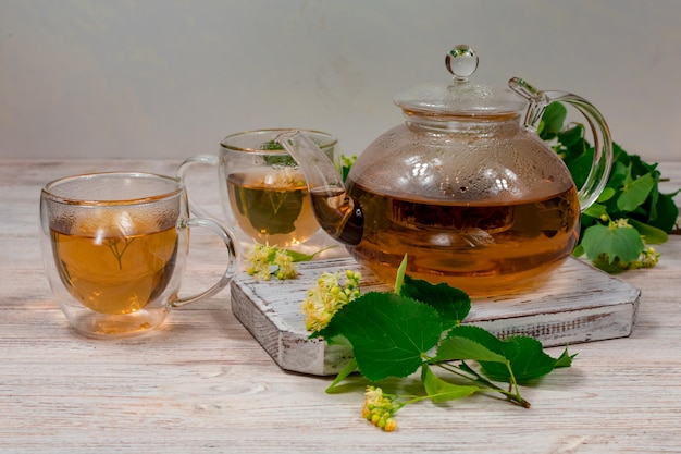 Szklany czajnik i dwie filiżanki herbaty z lipą na drewnianym stole na tle liści i kwiatów lipy Herbata lecznicza