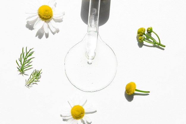 Szklany zakraplacz z produktem pielęgnacyjnym na bazie rumianku z kwiatami roślin na białym tle widok z góry