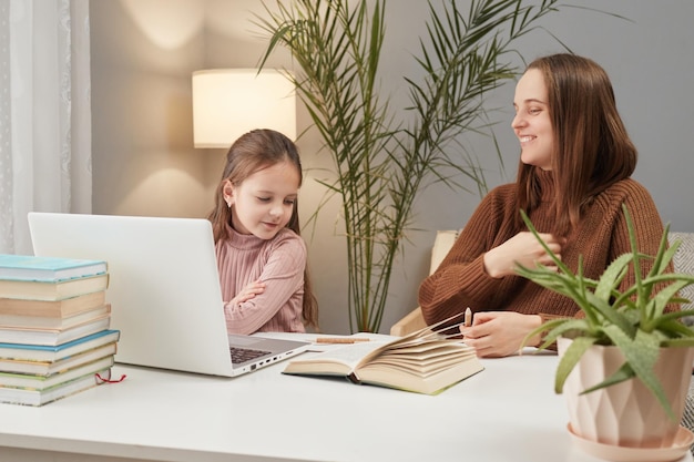 Szkolenie zdalne Szkolenie domowe Wesoła, radosna dziewczynka z nauczycielem używająca laptopa, siedząc przy stole w domu, ciesząc się wspólnym wykonywaniem zadań domowych