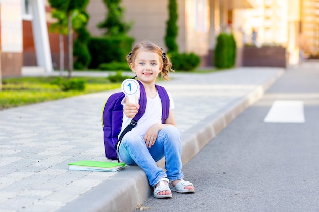 Szkolka z plecakiem i banknotami w rękach siedzi na chodniku w szkole i pokazuje numer jeden koncepcję powrotu do szkoły