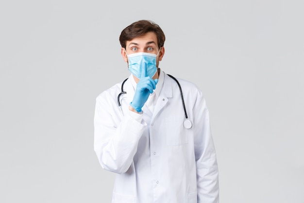 Szpital, pracownicy służby zdrowia, koncepcja leczenia Covid-19. Lekarz proszący zachować tajemnicę, milczeć, ma na sobie maskę medyczną, rękawiczki i peelingi, ucisza się z palcem przyciśniętym do ust.