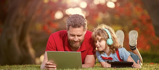 Sztandar ojca na syna szkoła chłopiec z laptopem nauka online leżąc na trawie mały chłopiec słucha muzyki w słuchawkach seminarium wideo lekcja online edukacja na laptopie