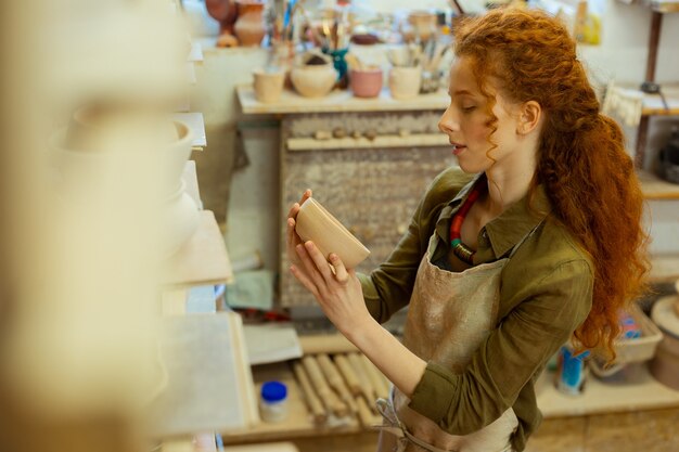 Szukać inspiracji. Zadowolona długowłosa mistrzyni garncarstwa obserwująca jej pierwsze dzieła, przebywając w pobliżu kolekcji ceramiki