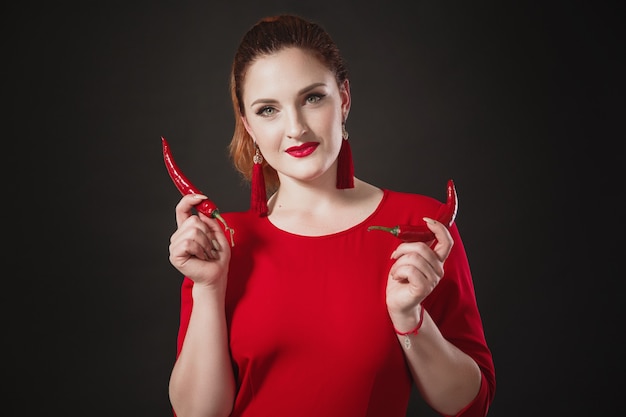 Szykowna rudowłosa młoda kobieta w czerwonym kombinezonie trzyma w dłoniach papryczkę chili Jasny wygląd.