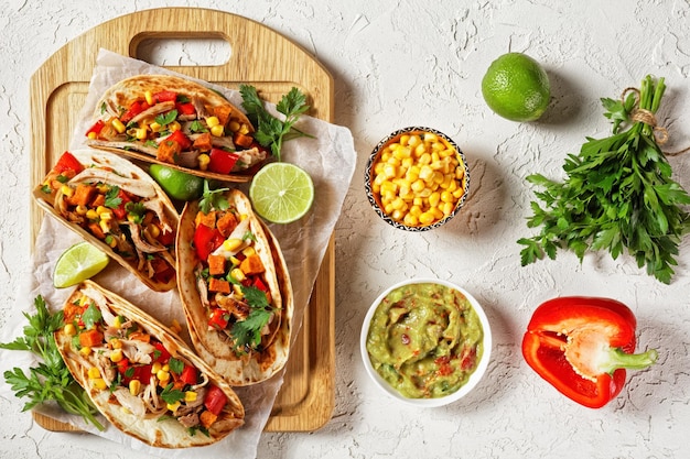 tacos z grillowanym mięsem z kurczaka, kukurydzą, pieczonymi kostkami batatów, czerwoną papryką i pietruszką podane na drewnianej desce na białym betonowym stole