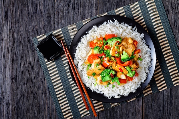 Tajski kurczak z orzechami nerkowca z warzywami stir-fry: brokułami, słodką papryką, ostrym czerwonym chili i sosem sojowym podany na czarnym talerzu z ryżem i pałeczkami na ciemnej drewnianej powierzchni, widok z góry, zbliżenie