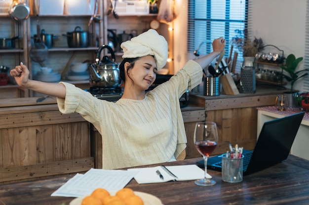 Tajwańska freelancerka z ręcznikiem na głowie wyciąga ręce podczas przerwy po skończonej pracy na laptopie przy stole w jadalni w domu.