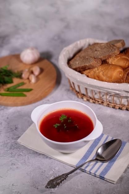 Talerz z barszczem buraczanym na stole, obok deski to pietruszka, koperek, zielona cebula i czosnek oraz kosz z chlebem. Smaczny i obfity lunch.