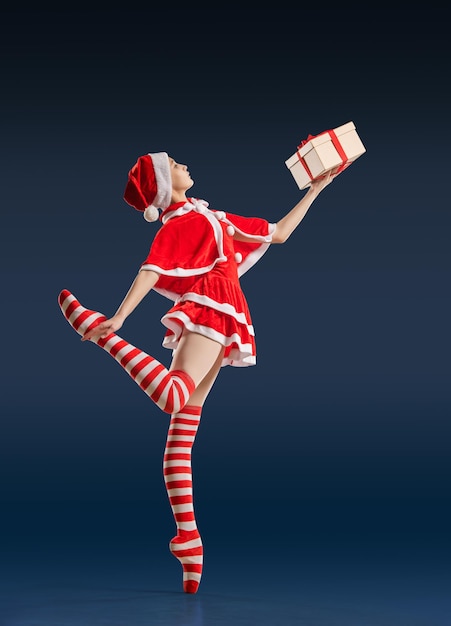 Tańcząca baletnica z prezentem w dłoniach na butach pointe w kostiumie Świętego Mikołaja na ciemnoniebieskim tle