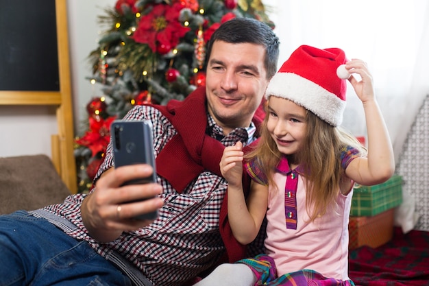 Tata I Córka Przy Choince Ze Smartfonem Robią Selfie, Komunikują Się Przez Połączenie Wideo. życzenia świąteczne, Pudełko, Dziewczynka W Czapce Mikołaja Machająca Hello.