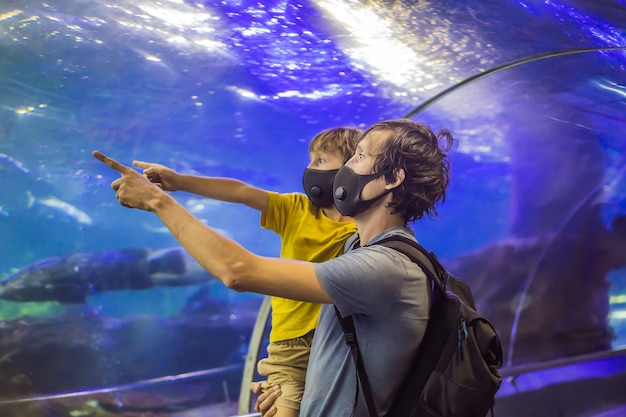 Tata i syn w maskach medycznych patrzą na ryby w akwarium w oceanarium, ludzie chodzą po medycynie