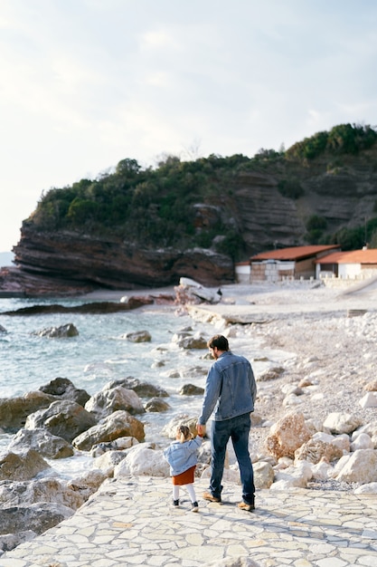 Tata prowadzi za rękę małą dziewczynkę po kamienistej ścieżce na kamienistej plaży do wody na tle