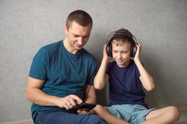 Tata siedzi na podłodze obok syna ze słuchawkami na uszach i wskazuje na telefon