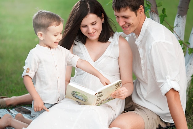 Tata syn i mama w ciąży patrzą na książkę w naturze pod drzewem i uśmiechają się