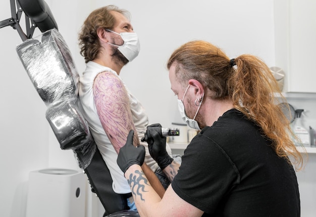 Tattooist ze swoją maszyną tatuuje na ramieniu z narysowanym projektem i używa maski ochronnej przeciwko covid 19 koncepcja sztuki i projektowania podczas nowej normalnej fotografii wysokiej jakości