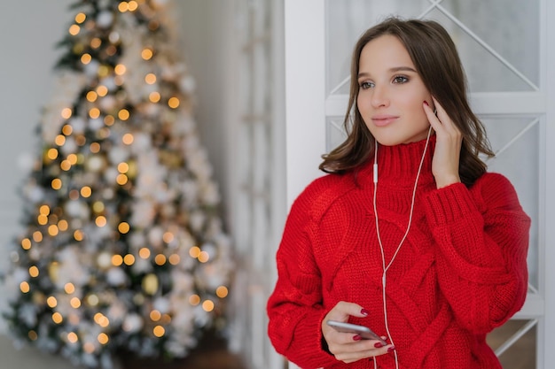 Techniczna kobieta cieszy się wakacjami, używa telefonu i słuchawek w pobliżu choinki.