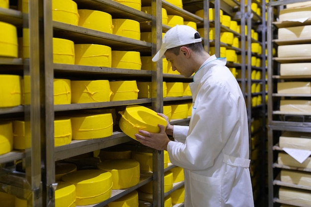 Technolog W Białej Szacie Z żółtą Głową Sera W Dłoniach Jest W Sklepie Do Produkcji Masła I Sera Proces Produkcji W Zakładzie Produktów Mlecznych Regały Z Serem